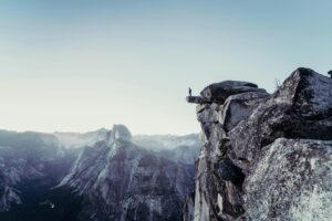 A man standing on the edge of a cliff in the mountains, overlooking a river flowing between peaks. The scene features hues of grey and blue, capturing the serene and dramatic landscape