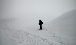 A man hiking in the tundra, capturing a photo of snowy hills and surrounding mountains. The landscape features stark white and black contrast, emphasizing the dramatic scenery.